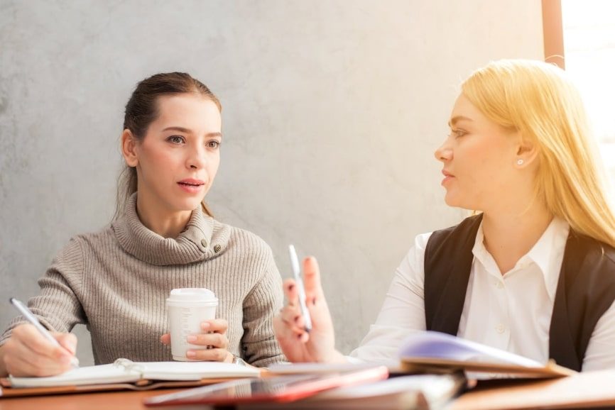 Two women having a conversation at a desk and communicating at work.